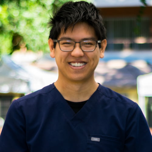 Alex Do, a smiling person wearing glasses and blue scrubs, stands outdoors.
