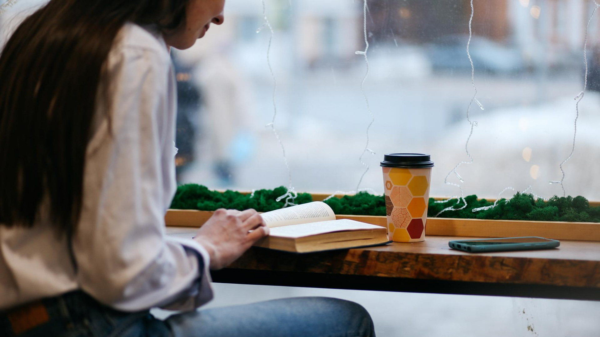 A person reading a book at a cafe table with a coffee cup and smartphone beside them.