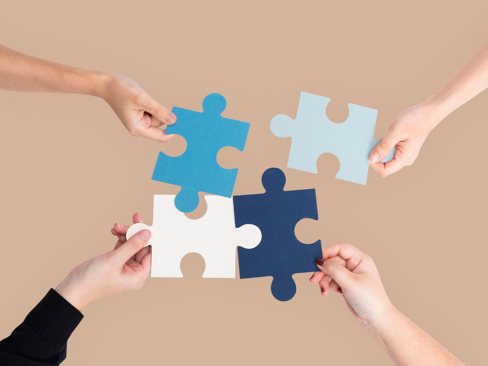 Four hands holding and connecting jigsaw puzzle pieces against a neutral background.