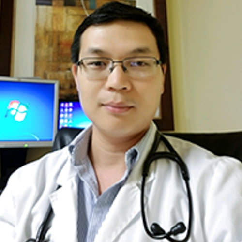 A man wearing glasses, a lab coat, and a stethoscope, identified as Thanh Nguyen NP-C, is sitting in an office with a computer monitor in the background displaying the windows desktop.
