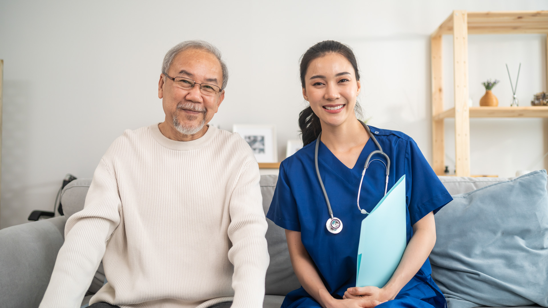 A smiling elderly man sitting beside a female nurse with a clipboard in a home setting.